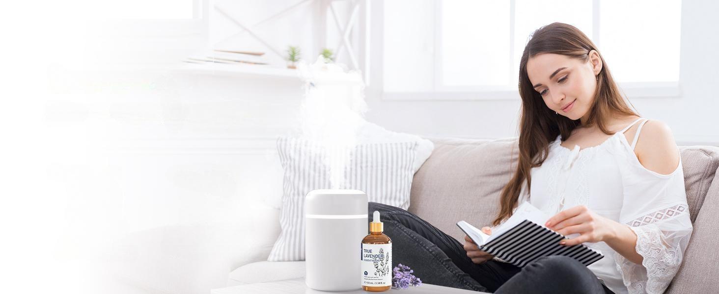 Young woman relaxing on a sofa reading a book while a white diffuser mists pure lavender essential oil to create a cozy, calming space in the living room.