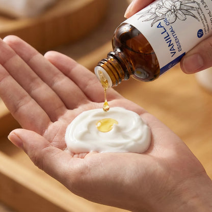 Close-up macro shot of a single golden drop of Vanilla Essential Oil being added to a dollop of rich white body lotion in a hand to create a DIY moisturizing blend.