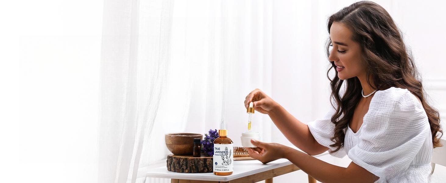 Smiling woman adding drops of pure lavender essential oil to her daily face moisturizer for a hydrating, soothing, and anti-aging skincare routine.